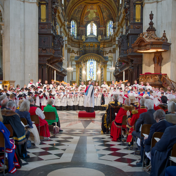 The Choir of St Paul's Cathedral mid-performance. A seated audience are watching. The floor is chequered, and the choir are dressed in white robes.