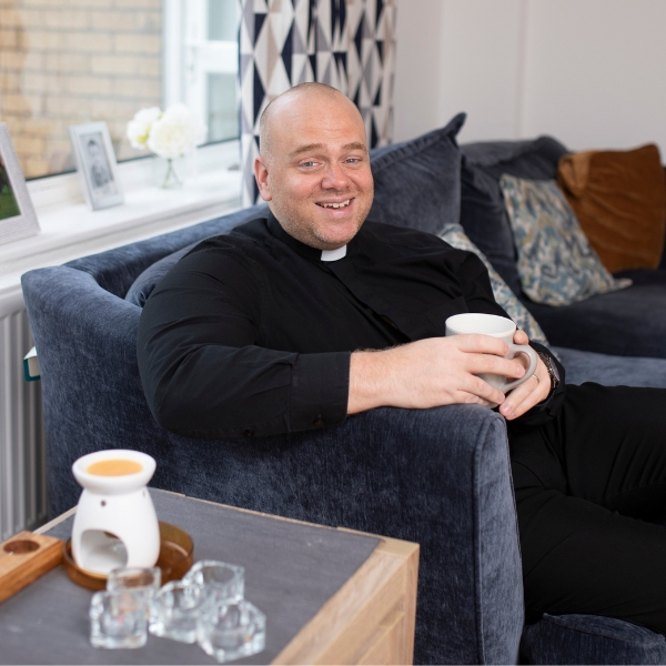 A man, dressed in black with a dog collar, drinking a cup of tea and smiling.
