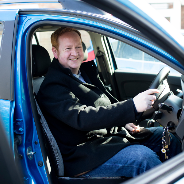 A man, sitting in the front seat of his car, smiling and looking at the camera.