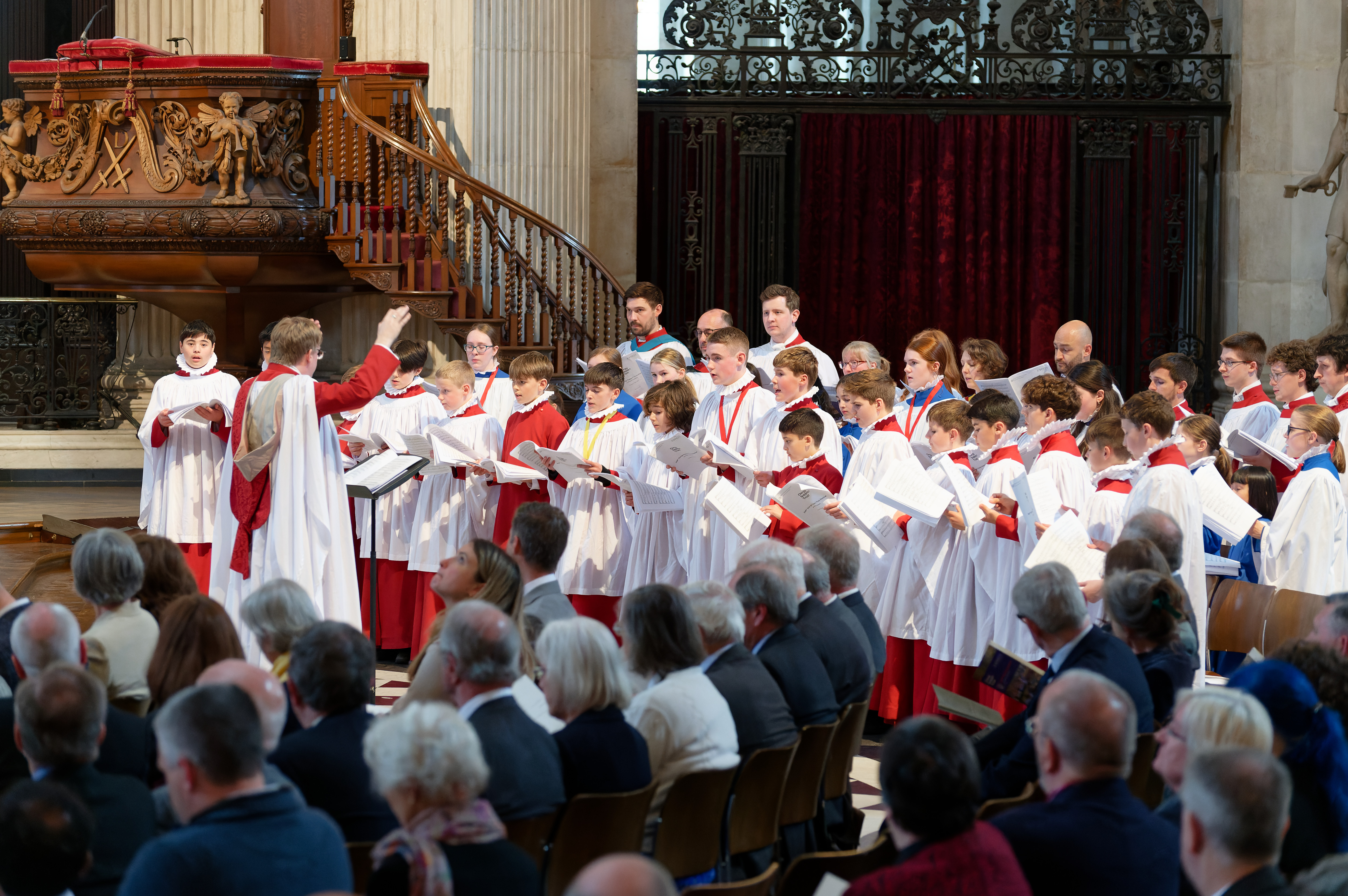 A choir of young people, dressed in red and white robes, performing, led by a choir master to a seated audience.