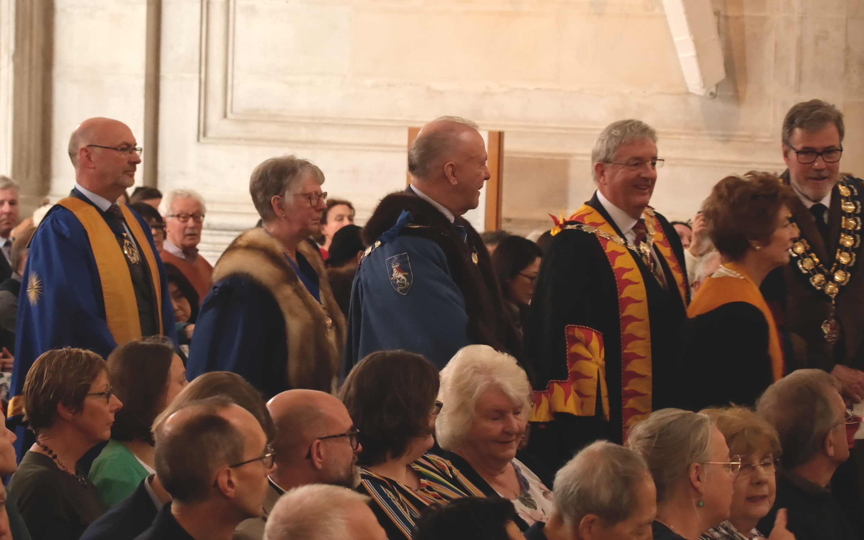 In the middle of a seated audience, five people walk forward, dressed in blue, yellow and gold gowns.