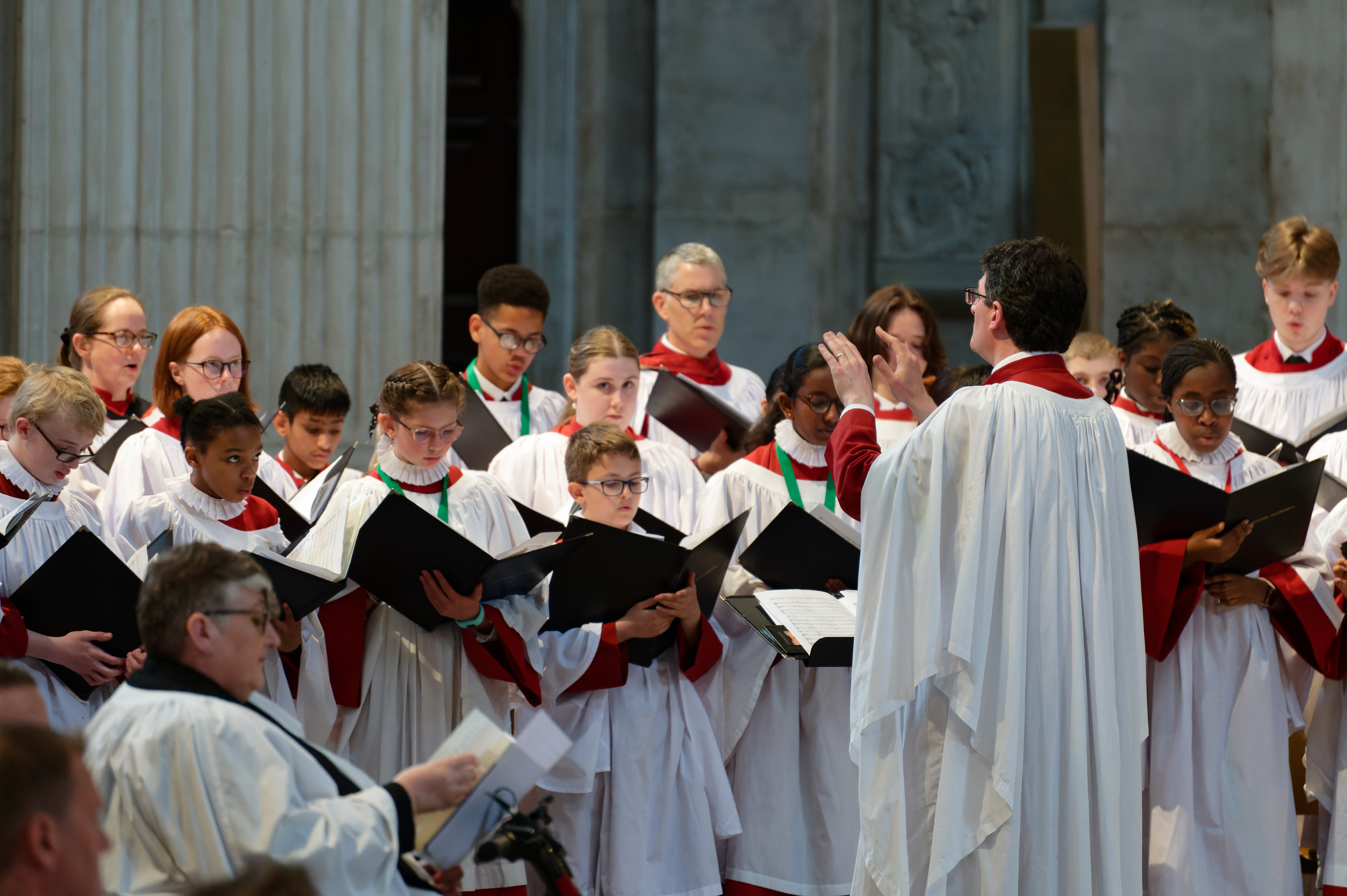 Led by a choirmaster with black hair and glasses, a choir of young people in white robes and green lanyards sing from their songbooks.