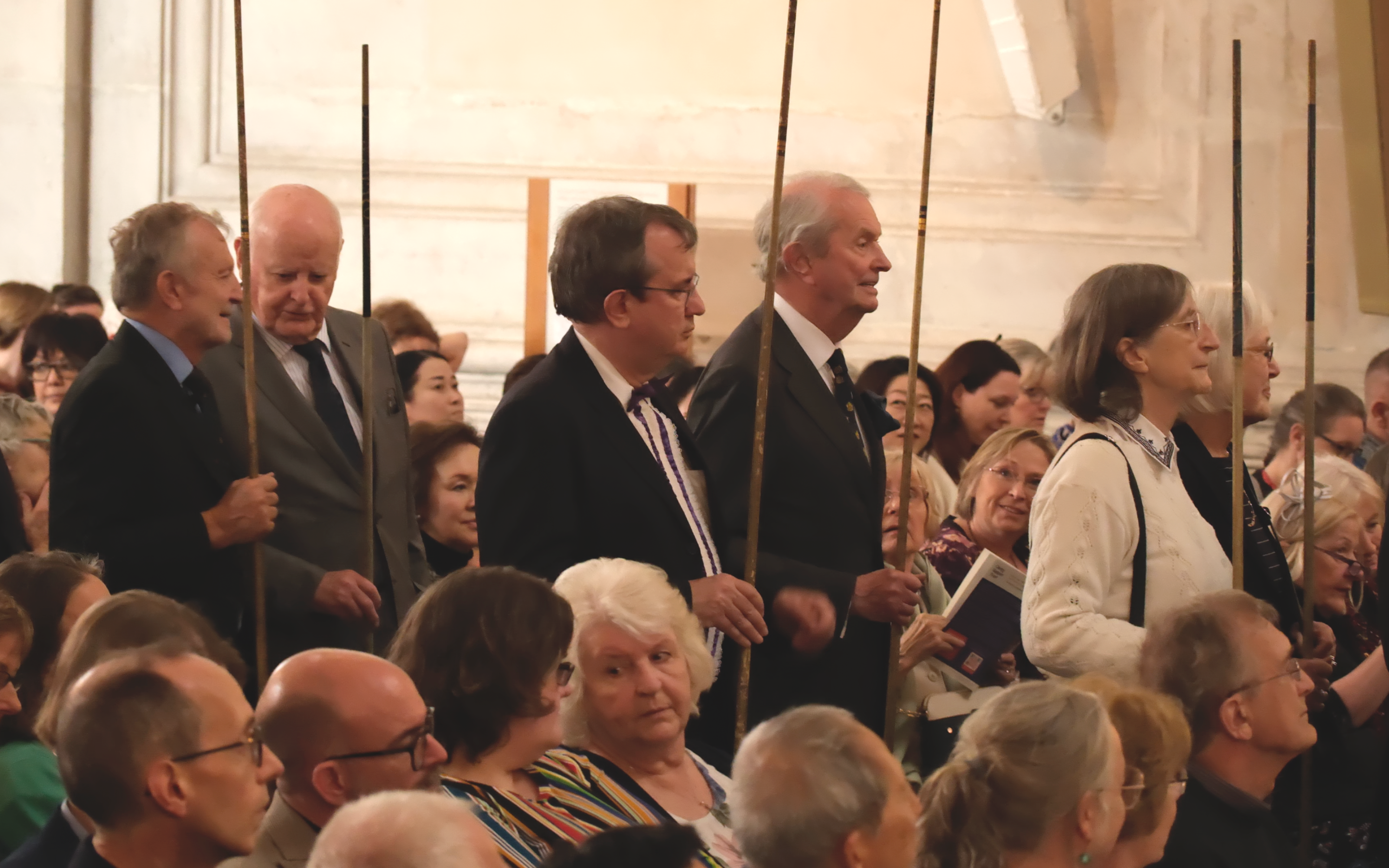 In the middle of a seated audience, six people walk forward, dressed smartly, carrying large ceremonial sticks.