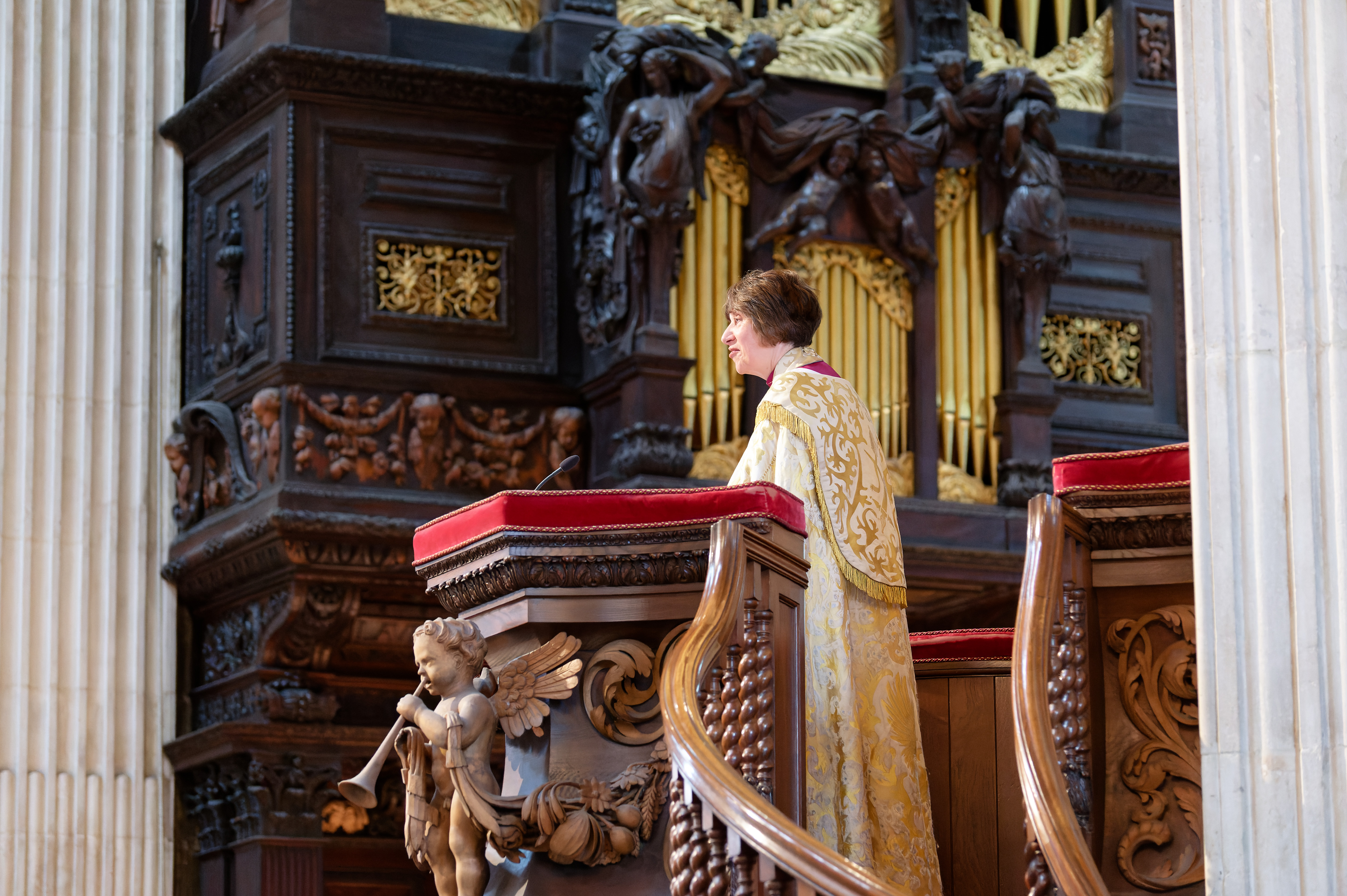 The Right Revd Rachel Treweek, dressed in a white and gold gown, speaking from a wooden pulpit. Behind her is a grand black and gold organ.