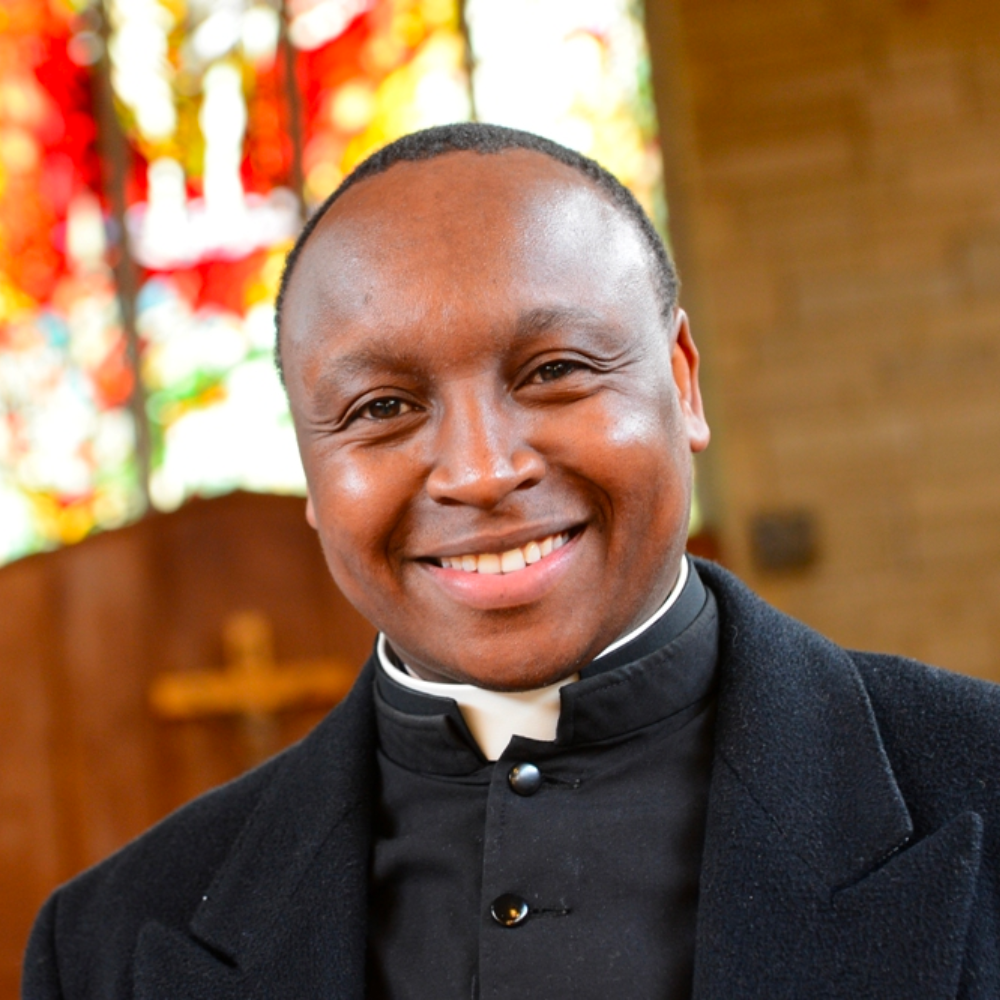 A vicar, dressed in black and standing in front of a stained glass church window.