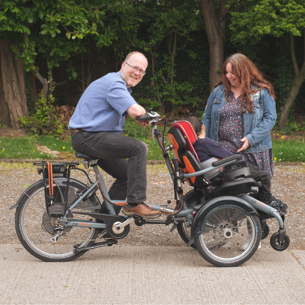 Mira on her new bike, with her father sat behind her and her mother standing in front. In the background is a woodland.