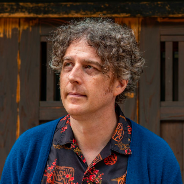 A man with curly hair, standing in front of a dark brown wooden gate.