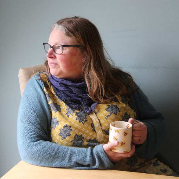 Becca, wearing a light blue cardigan. She is sat at a table, drinking a cup of tea, looking out of the window.