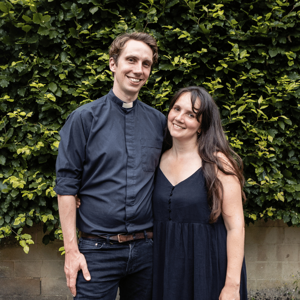 Matt is wearing a grey shirt and a white clerical collar. Esther is wearing a black dress. The two are holding one another, while looking at the camera and smiling. They are stood in front of a garden hedge.