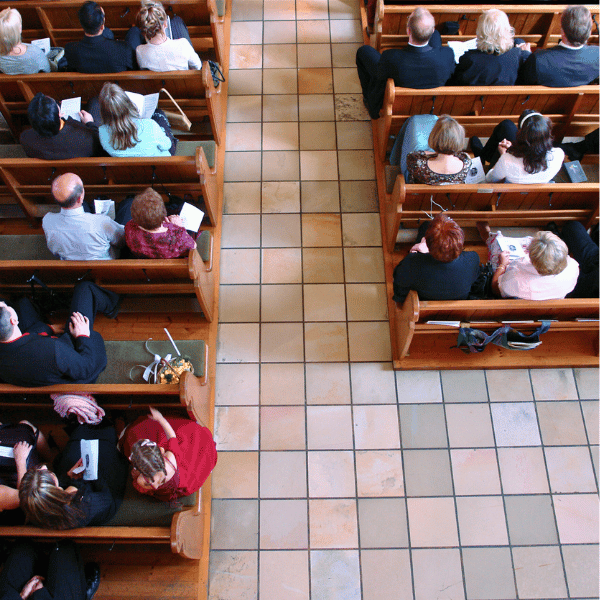An aerial short of congregants sat on wooden pews in church, upon a tiled floor.