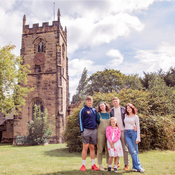 The family are stood together, looking at the camera. Behind them are a church tower, tall trees and a cloudy sky.