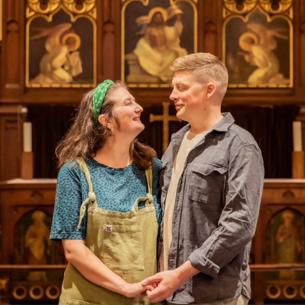Becca is dressed in a blue t-shirt and dungarees. James is dressed in a white t-shirt and a dark grey shirt. Both are holding hands, and looking into one another's eyes.