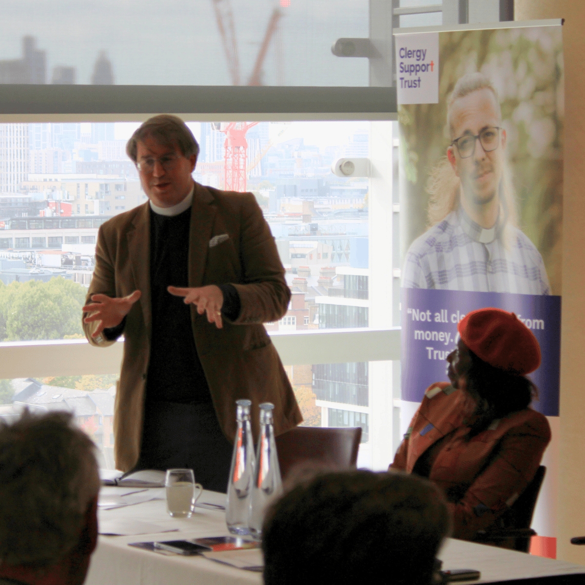 Our Chief Executive, The Revd Ben Cahill-Nicholls, is dressed in a black shirt, white clerical collar and a brown jacket. Behind him is a view from the window of the London skyline.