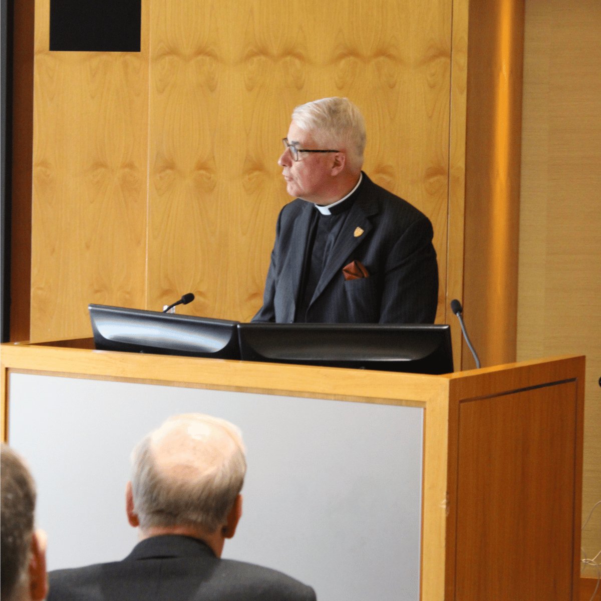 The Revd Canon Thomas Woodhouse, dressed in a black shirt, black jacket and white clerical collar, is speaking from behind a wooden lectern.