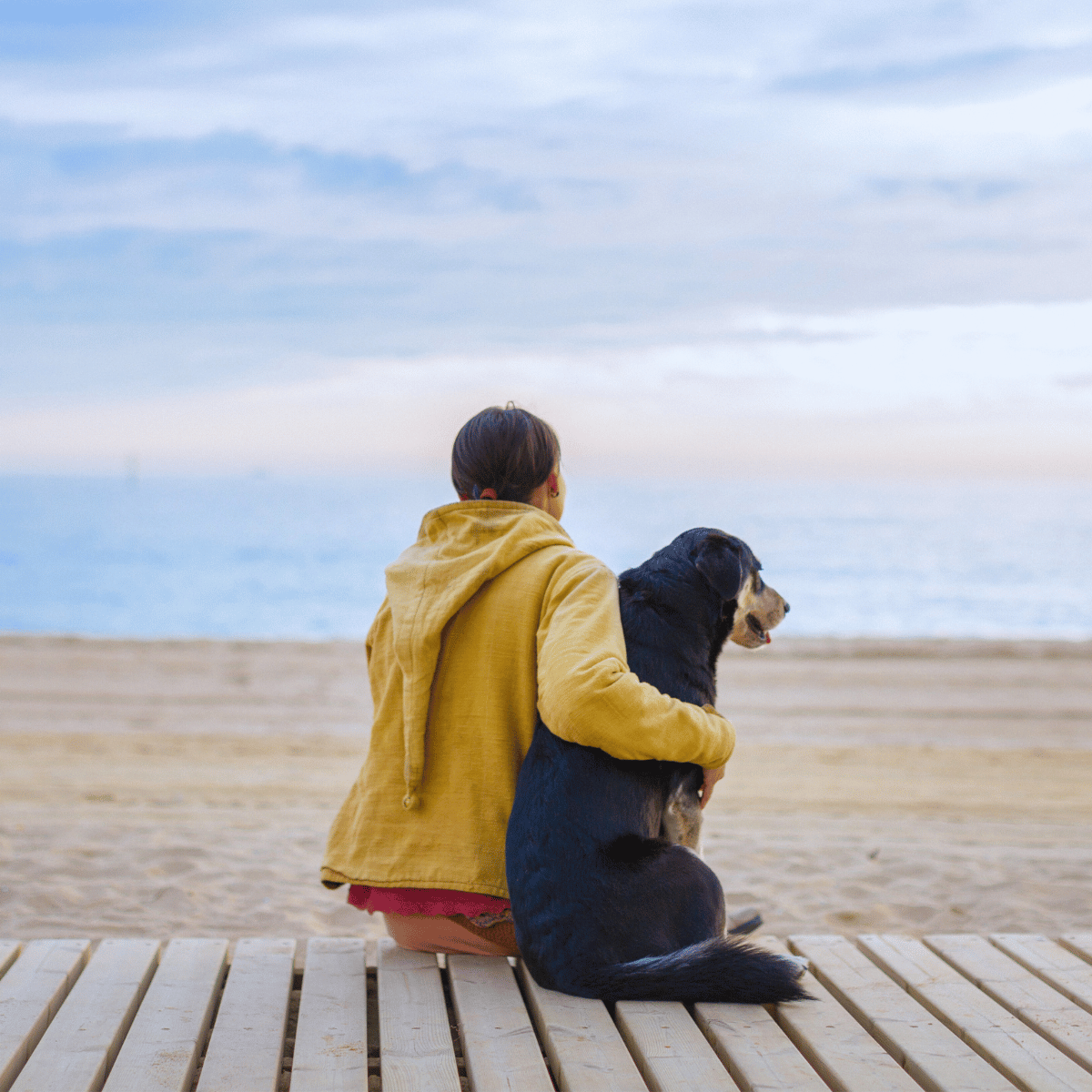 A woman, dressed in a yellow raincoat, is sat on the beach, cuddling her dog.
