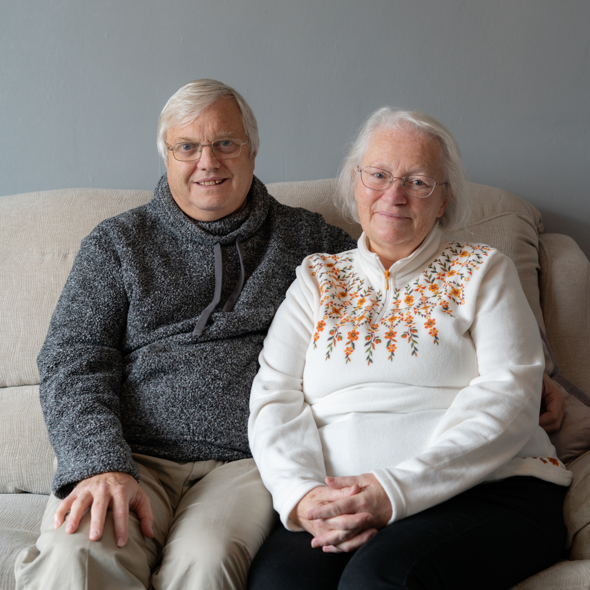 Paul is dressed in a grey hoodie, while Jo is wearing a white jumper. They are both sat on their sofa in the living room.
