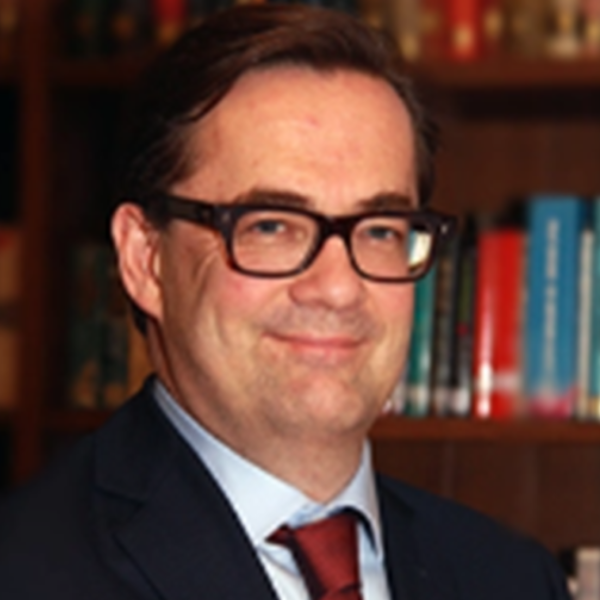 Peter Warren is wearing glasses and a formal suit, and is stood in front of a bookcase.