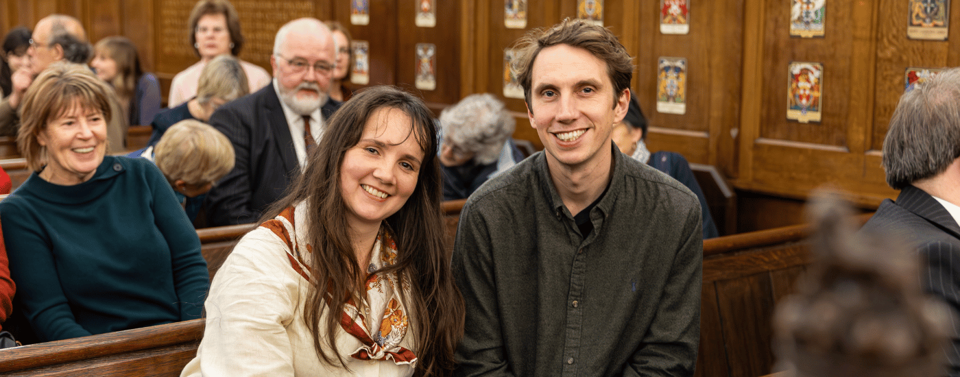 A man and a woman are sat on a wooden pew, smiling at the camera.