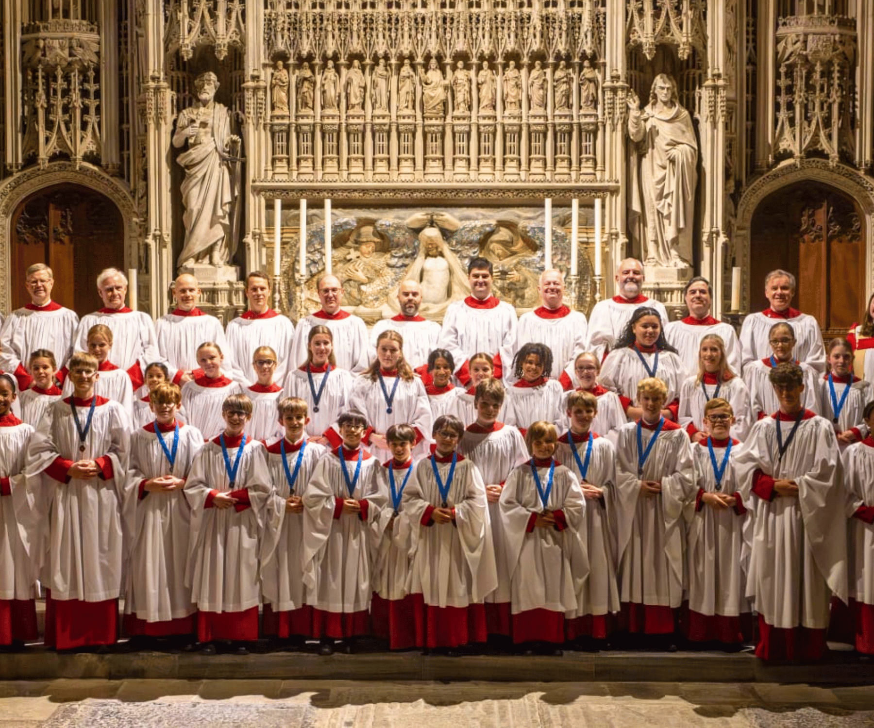 Choristers in white and red robes are stood in three lines, smiling and looking at the camera. Behind them are stone statues of saints and religious figures.