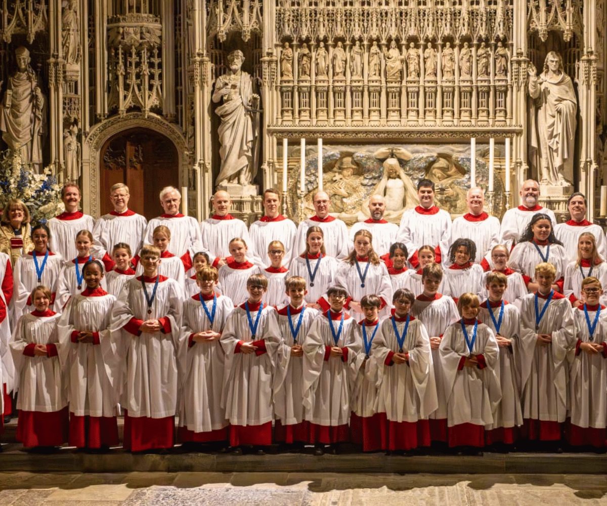 Three lines of adult and child choristers are dressed in white robes, looking at the camera and smiling.