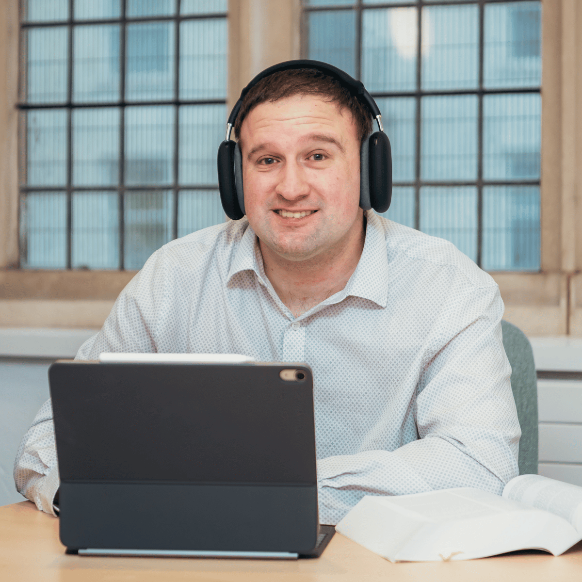 Ben is sat at a desk. He is wearing a white shirt, headphones and using an iPad.
