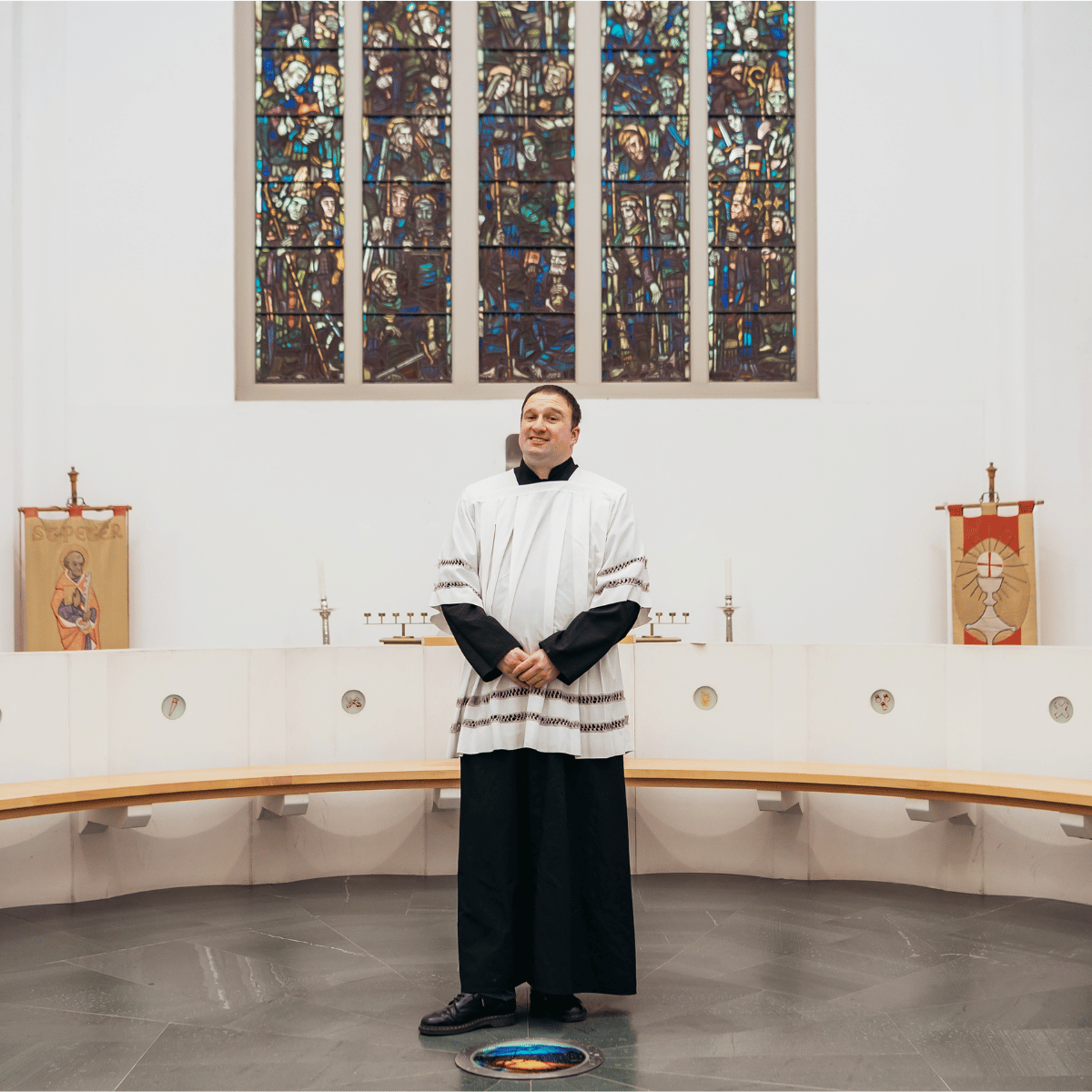 Ben is wearing a white robe, and is standing in front of a stained glass Church window.