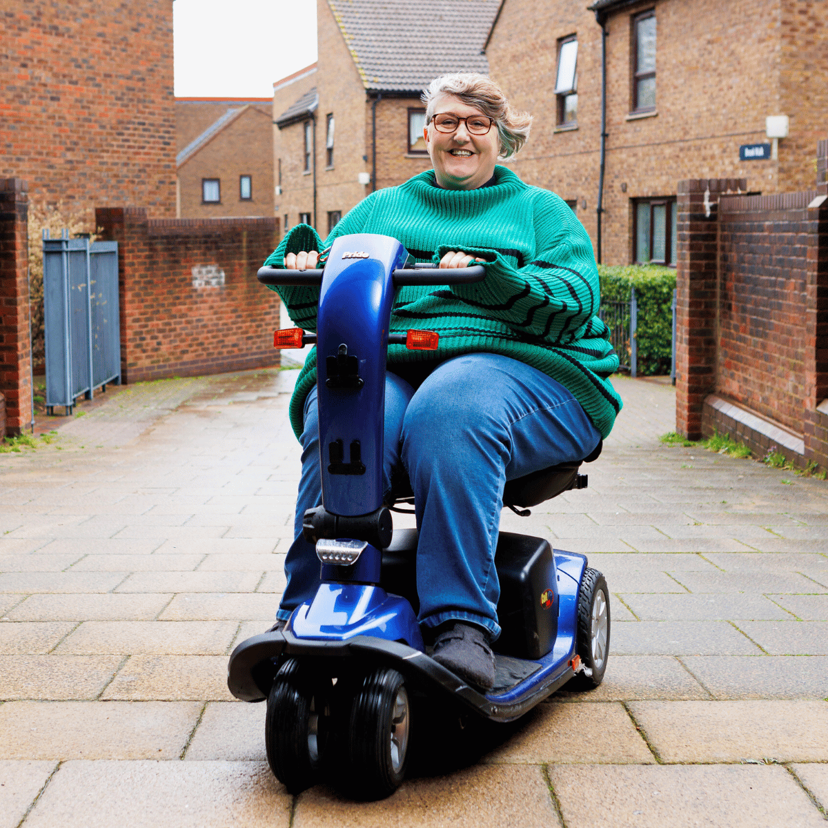 The Revd Helen Shannon, wearing a green jumper and navy blue jeans, is using an electric mobility scooter on a pavemented side street.