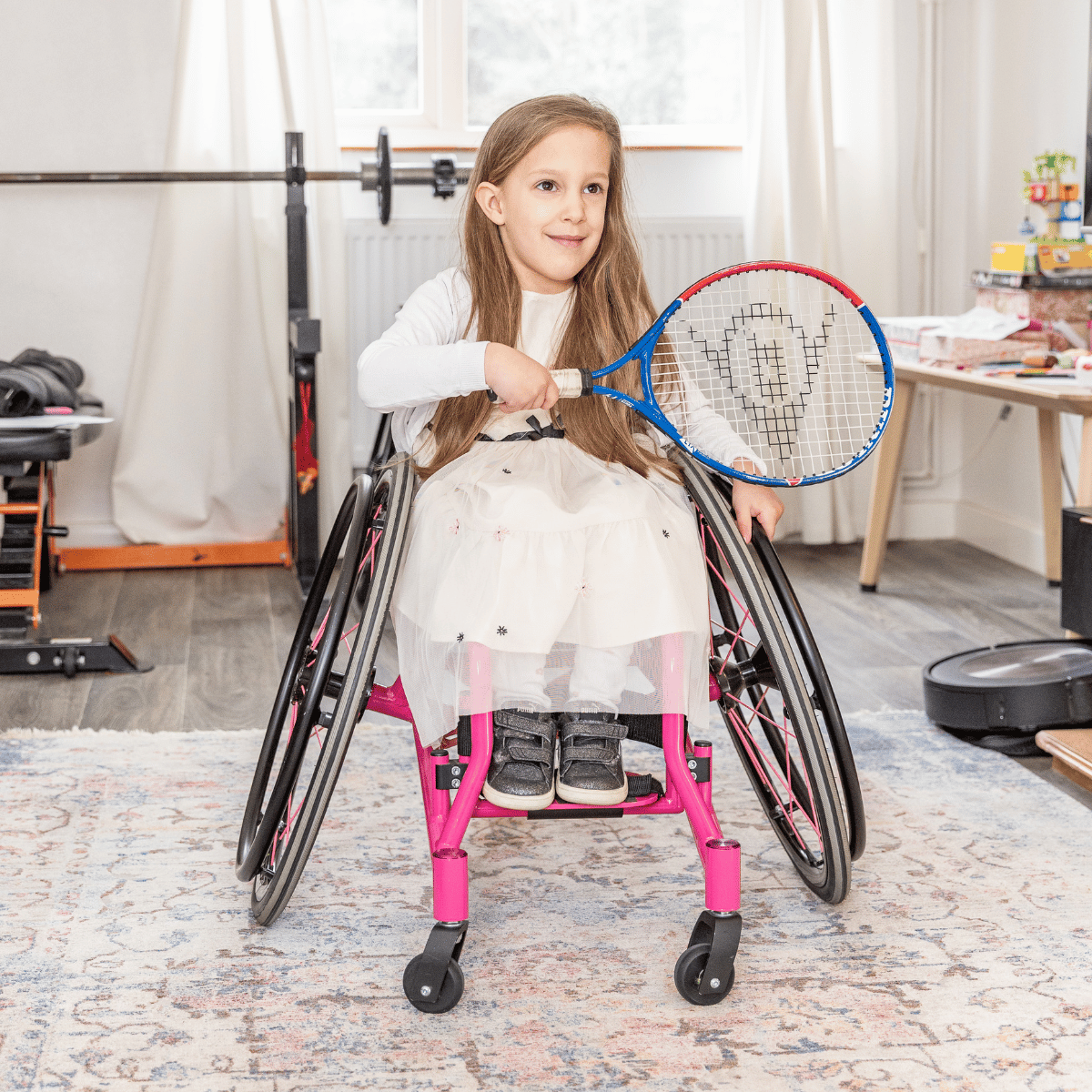 Lina is in a white dress, sat in her wheelchair, holding a tennis racket, in the living room.