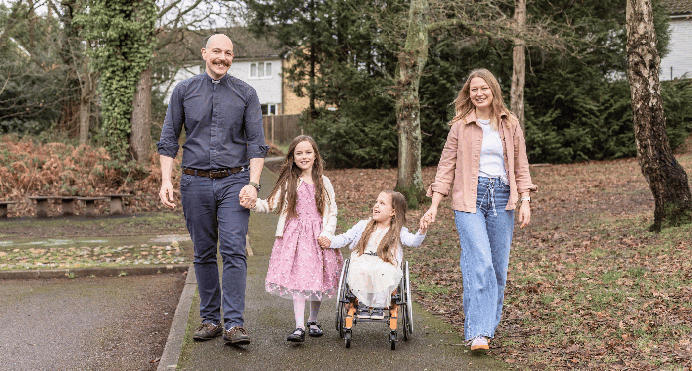Nik, Heidi and their daughters are outside, in an autumnal setting, with brown trees and bushes behind them. They are holding hands.