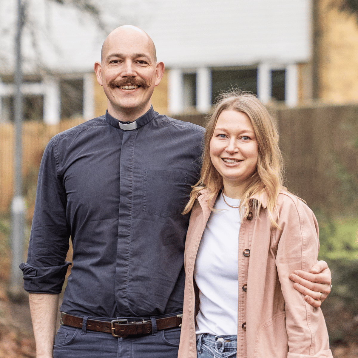 Nik is wearing a grey shirt and a white clerical collar. Heidi is wearing a white t-shirt and a salmon jacket. They are stood outside, and Nik has his arm around Heidi.