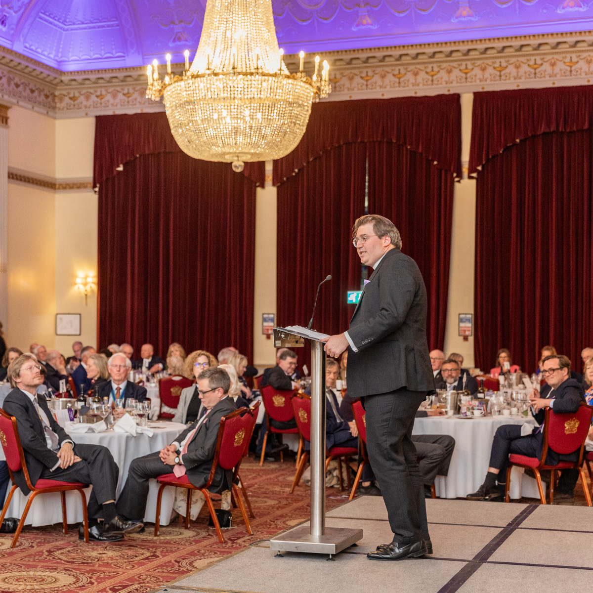 The Revd Ben Cahill-Nicholls is dressed in a black suit and standing behind a podium. He is addressing a room of seated guests. A grand, bright chandelier is hanging from the ceiling.