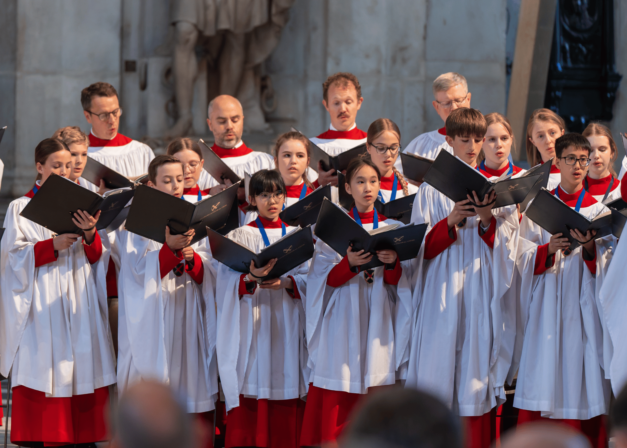 St Albans Cathedral Choir are dressed in white and red robes, singing together from their song books.