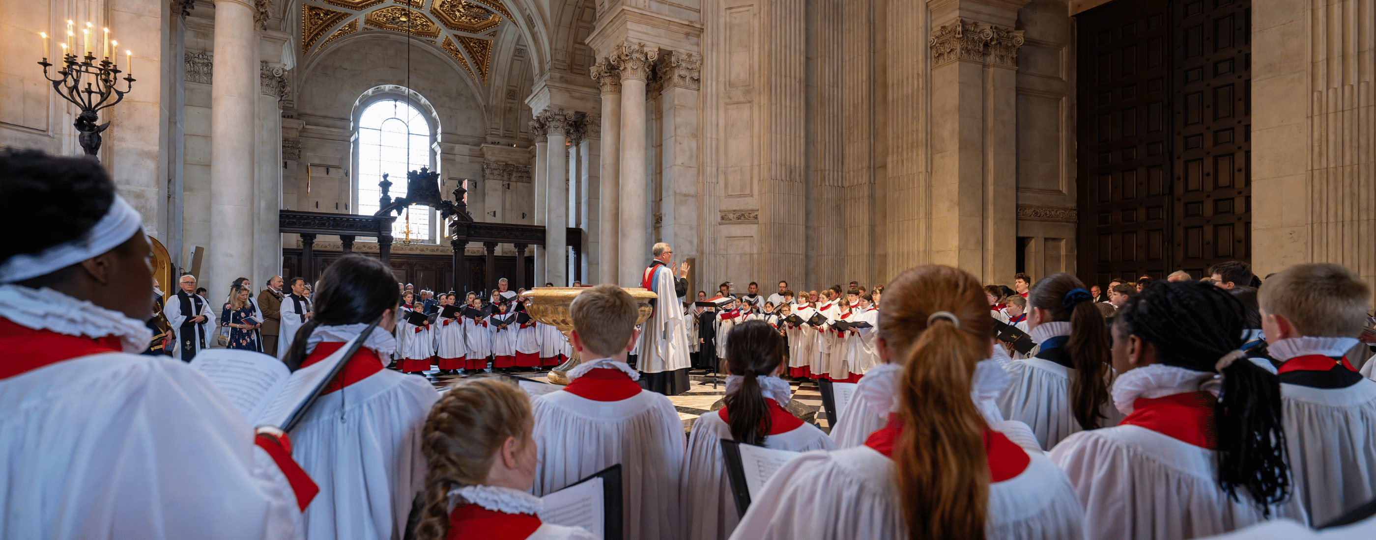 The choirs of Blackburn, St Albans and St Paul's Cathedral are dressed in white and red robes, and are singing together. They are led by Director of Music, Andrew Carwood MBE, and performing at the West Doors of St Paul's Catheral, surrounding the font.