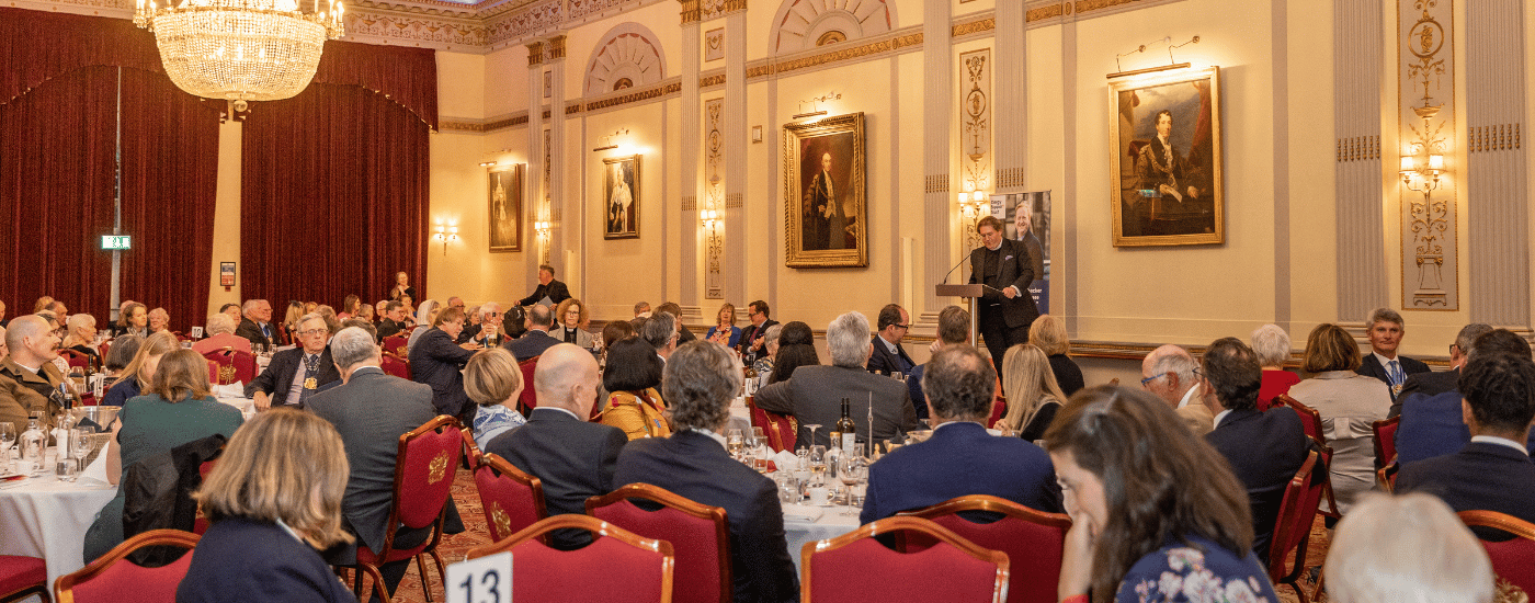 The interior of Plaisterers' Hall. Paintings of past Livery Masters are hung in golden frames. A grand chandelier hangs from the ceiling. Seated guests are listening to a speaker, stood on stage at a podium.