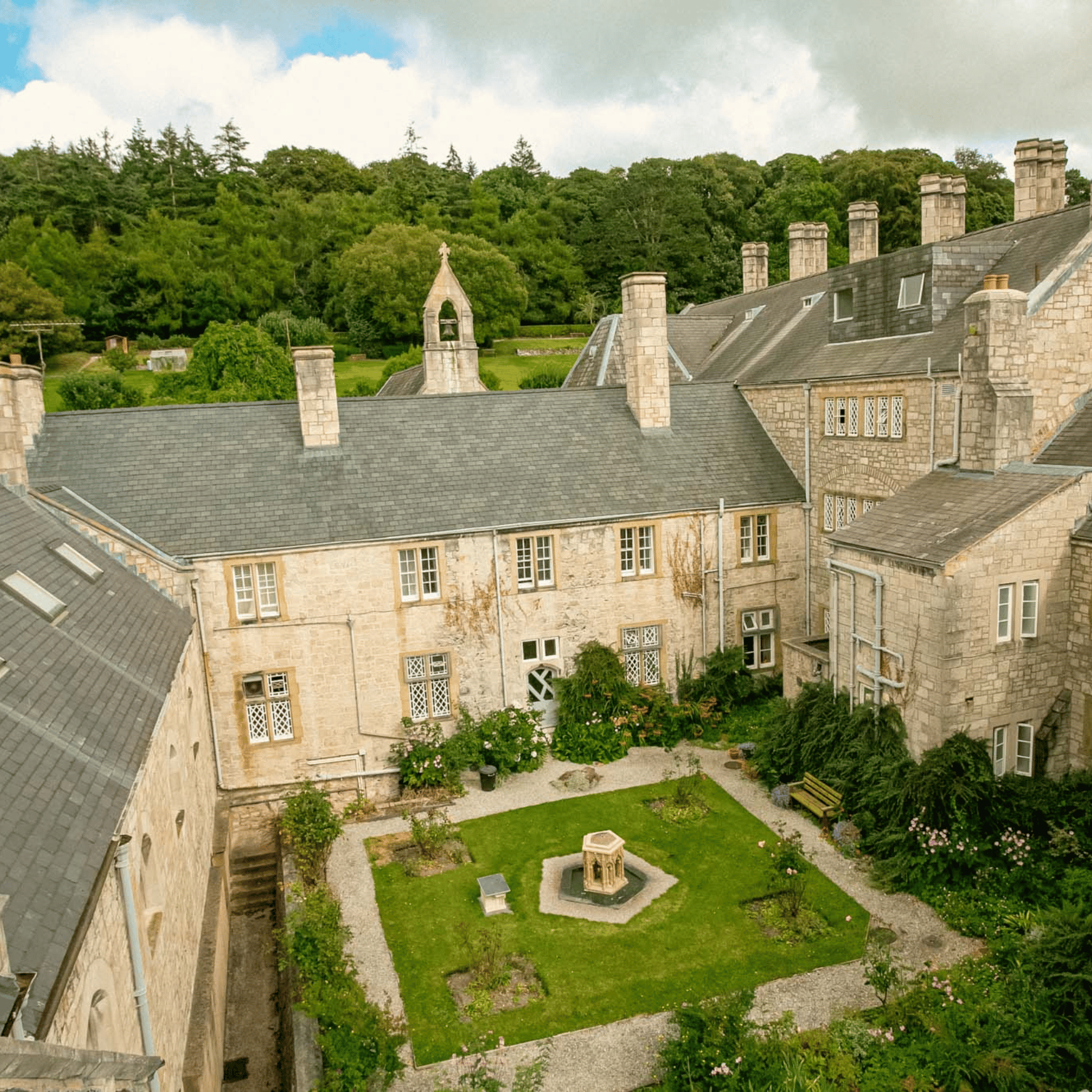 St Beuno's Jesuit Spirituality Centre. A courtyard with a water fountain, and a stone footpath circling a square lawn. Old cottages surround the courtyard on all sides. In the distance is a bright and vibrant woodland.
