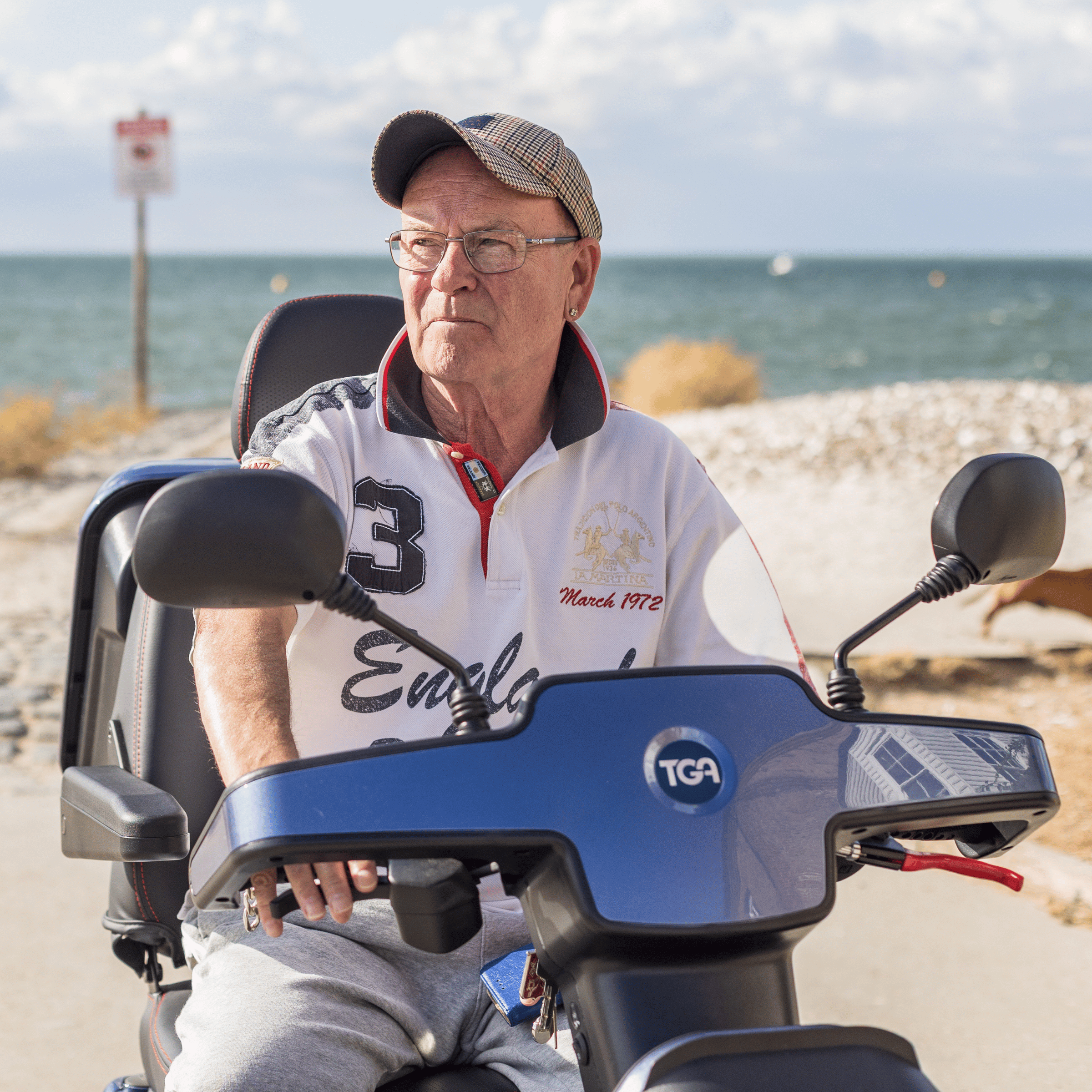 Martin is sat on a dark blue mobility scooter. Behind him is a long stretch of sandy beach, leading to the ocean.