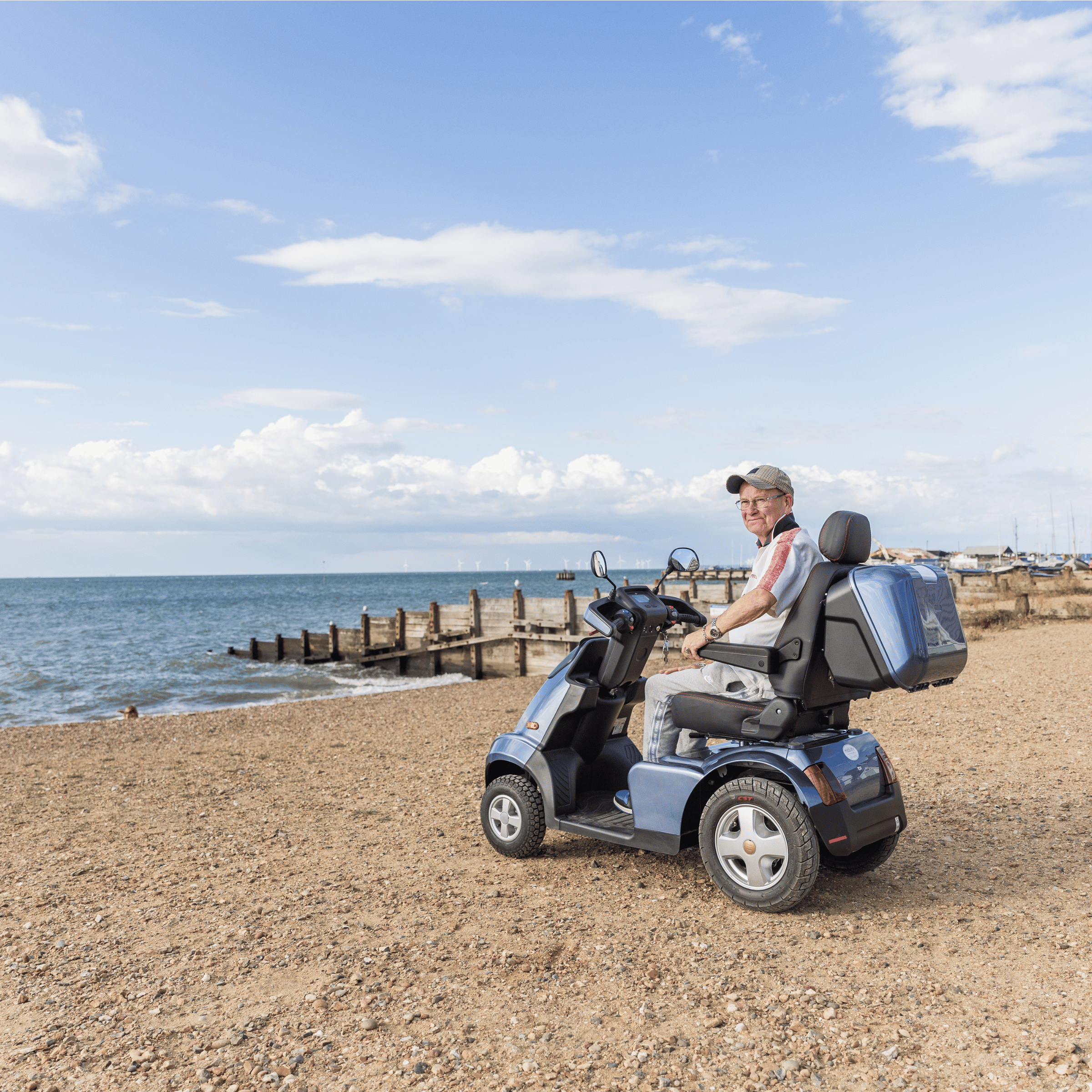 Martin is sat on a dark blue mobility scooter, parked on the beach, facing the sea. The sky is a tranquil blue, with light clouds.