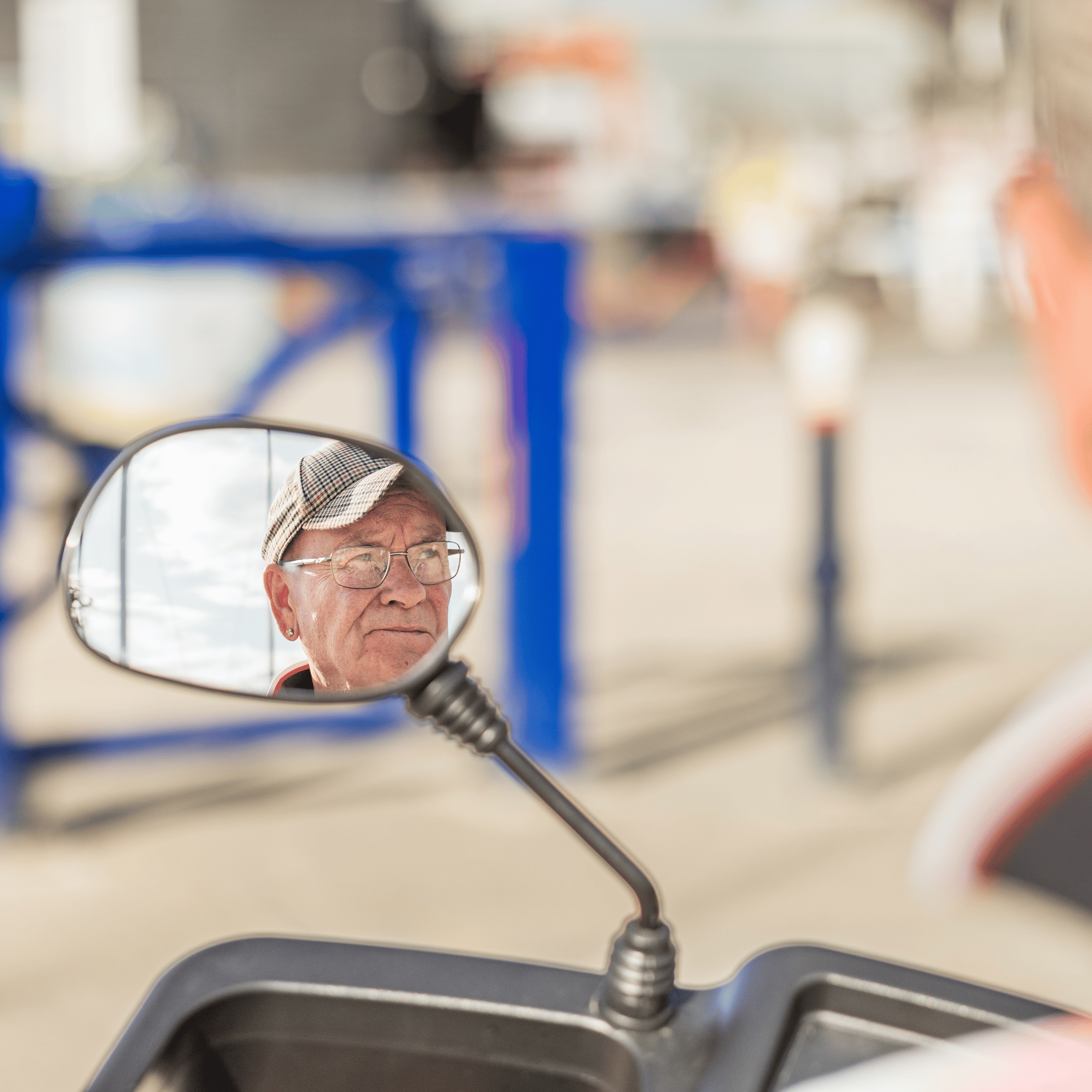 A zoomed in photo of the rear-view mirror of a mobility scooter. Martin is in the reflection, wearing glasses and a cap.