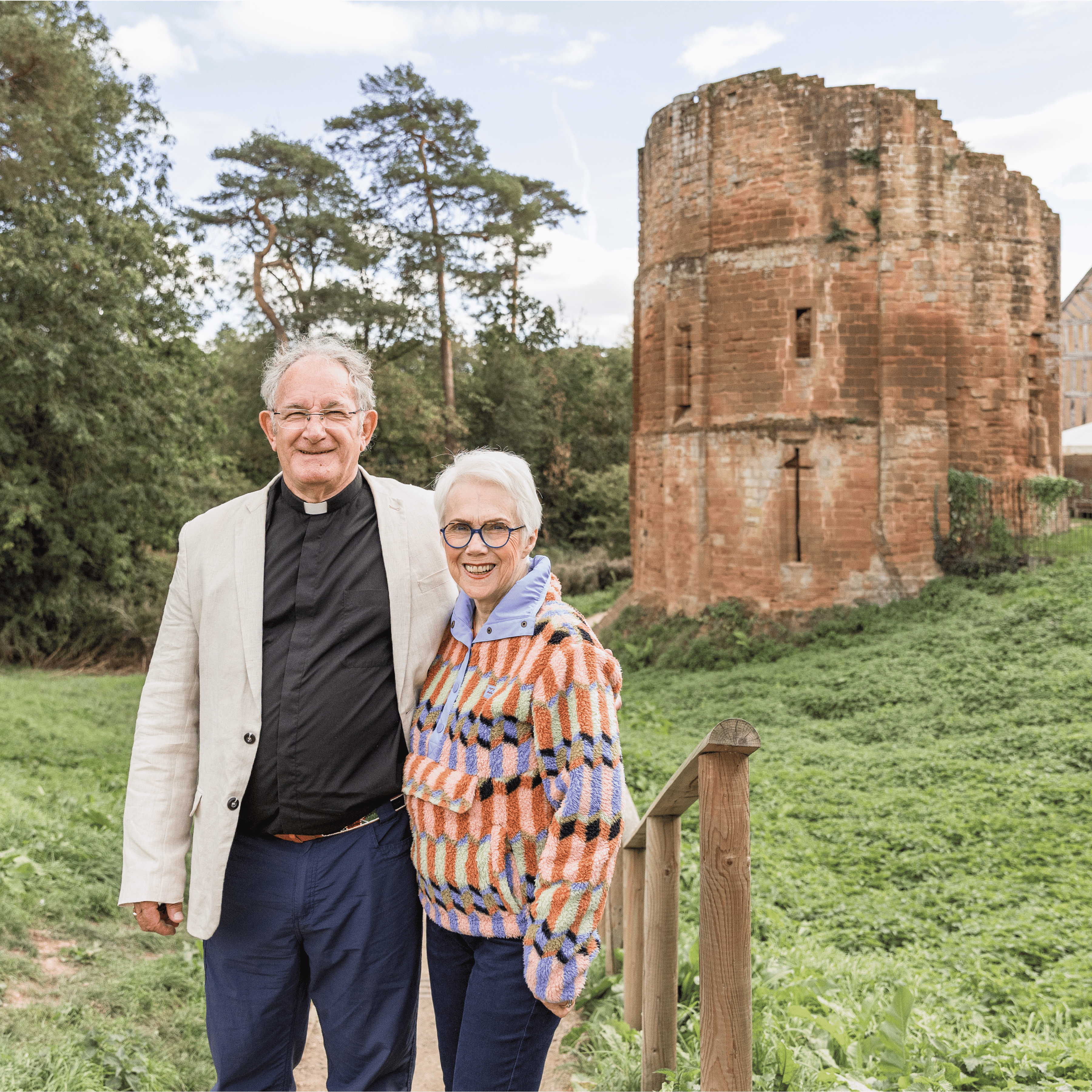 Michael and Sybil are posing in front of the remains of an abandoned stone building, an old chapel or castle, surrounded by overgrown grass.