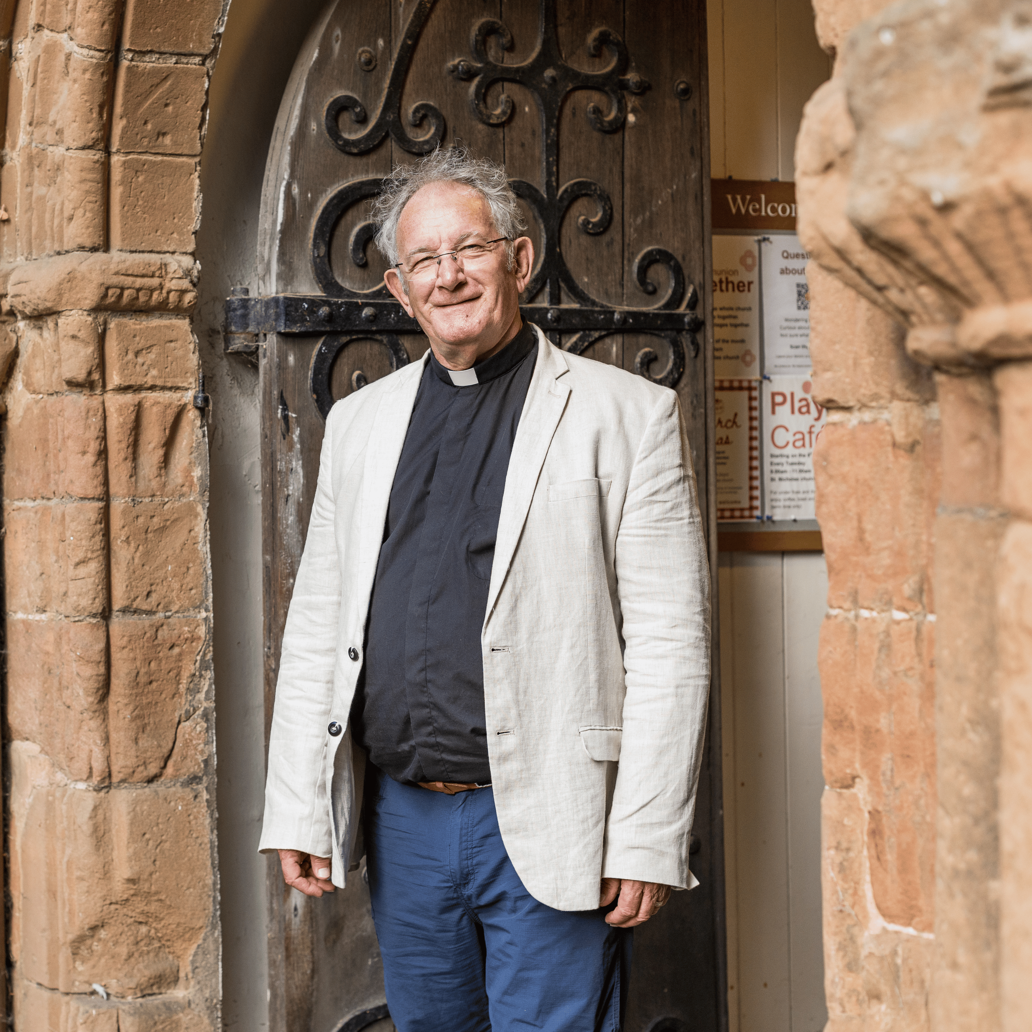 Michael is wearing a cream jacket, black shirt and white clerical collar. He is stood beside the old wooden doors of a church, beneath a stone archway.