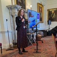 Catherine holding a microphone and standing in front of a TV screen to give a presentation.