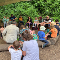 A group of children, sitting on logs in a circle, in front of a row of green trees and bushes.