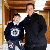 Josiah and his mother, Hannah. Both are dressed in black, facing the camera and smiling. Behind them is the orange-brown wood of the stables.