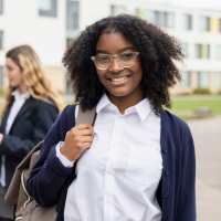 A little girl, standing outside a school. Two other students are standing behind her. She is wearing glasses, a white shirt and a blue jacket.