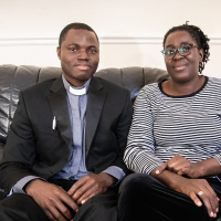 Joseph, in a black suit jacket, a blue shirt and a white dog collar, sitting on a leather sofa beside his wife, in glasses and a striped t-shirt.