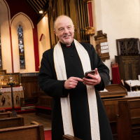 A vicar, wearing a black robe and white clerical collar, standing in his Church and using his mobile phone.