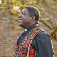 A vicar, wearing a black shirt and a white clerical collar, standing in front of a meadow of trees.