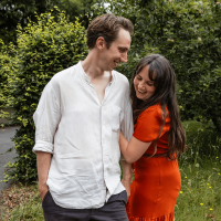 Matt is wearing a white shirt. Esther is wearing an orange dress. The two are looking at each other fondly, stood outside beside green grass and a tall hedge.