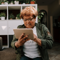 A woman, dressed in a grey and white striped shirt and dark green jacket, is using an iPad in her home.