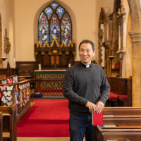 Jun is wearing a black shirt, white clerical collar, and holding a red hymn book, while standing in front of a stained glass window.